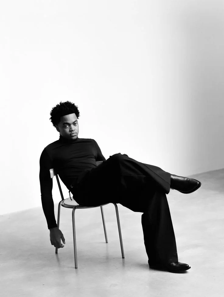 Black-and-white studio portrait of a man in black reclining on a metal chair.